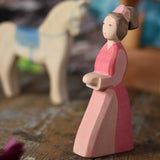An Ostheimer hand made wooden Lady-In-Waiting toy figure placed on a wooden surface in a small world play scene. Other wooden toys including an Ostheimer water well can be seen in the background. These wooden figures are part of a wide range of expertly hand crafted wooden toys here at Babipur. 