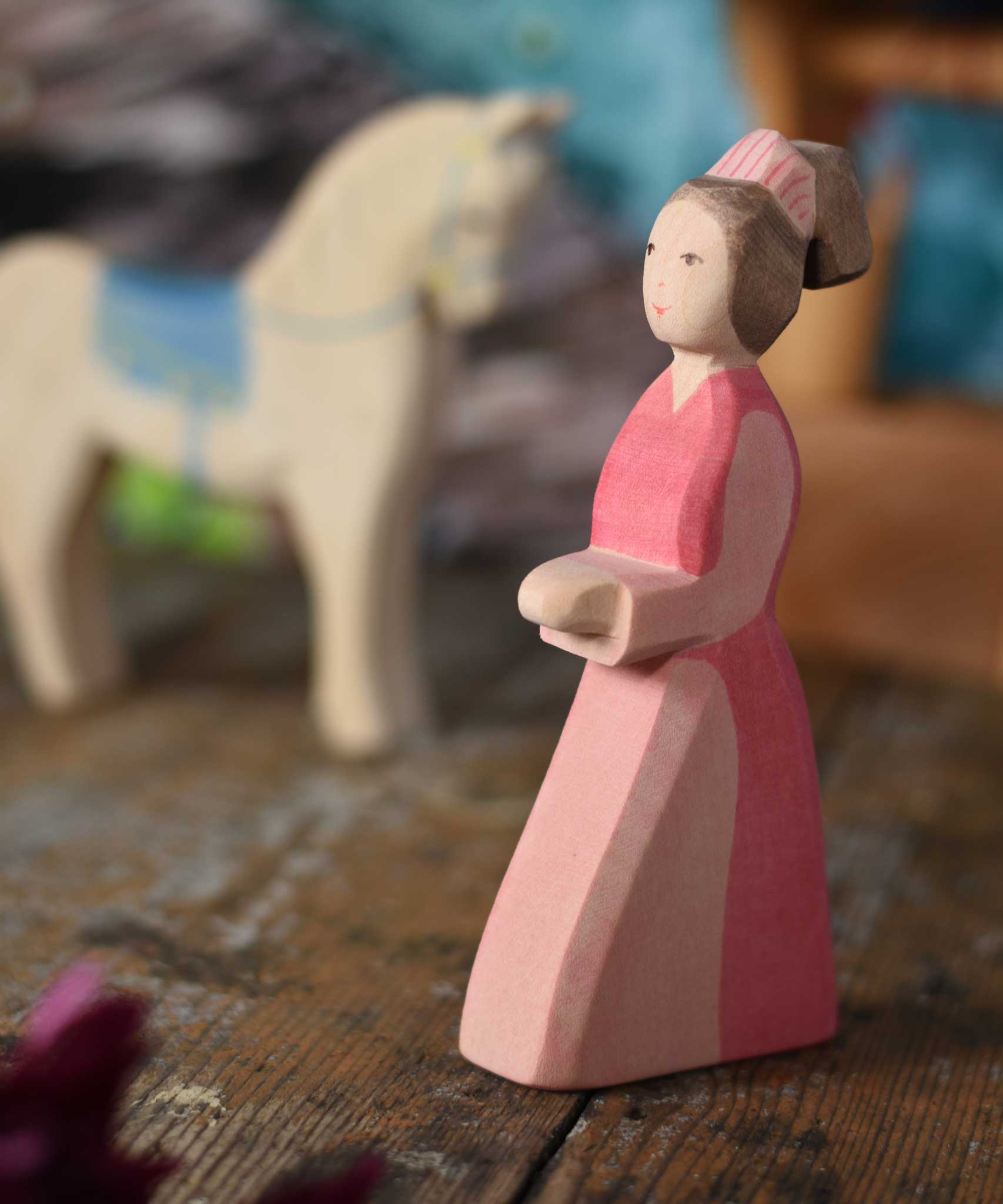 An Ostheimer hand made wooden Lady-In-Waiting toy figure placed on a wooden surface in a small world play scene. Other wooden toys including an Ostheimer water well can be seen in the background. These wooden figures are part of a wide range of expertly hand crafted wooden toys here at Babipur. 