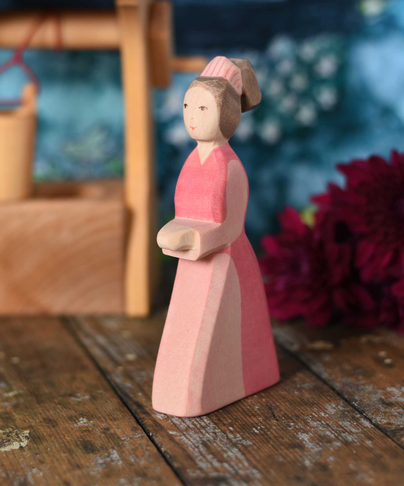 An Ostheimer hand made wooden Lady-In-Waiting toy figure placed on a wooden surface in a small world play scene. Other wooden toys including an Ostheimer water well can be seen in the background. These wooden figures are part of a wide range of expertly hand crafted wooden toys here at Babipur. 