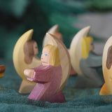 A purple coloured little Ostheimer angel placed on a green decoration scarf in a small world play set up. Other little angels can be seen in the background. These wooden figures are part of a wide range of expertly hand crafted wooden toys here at Babipur.