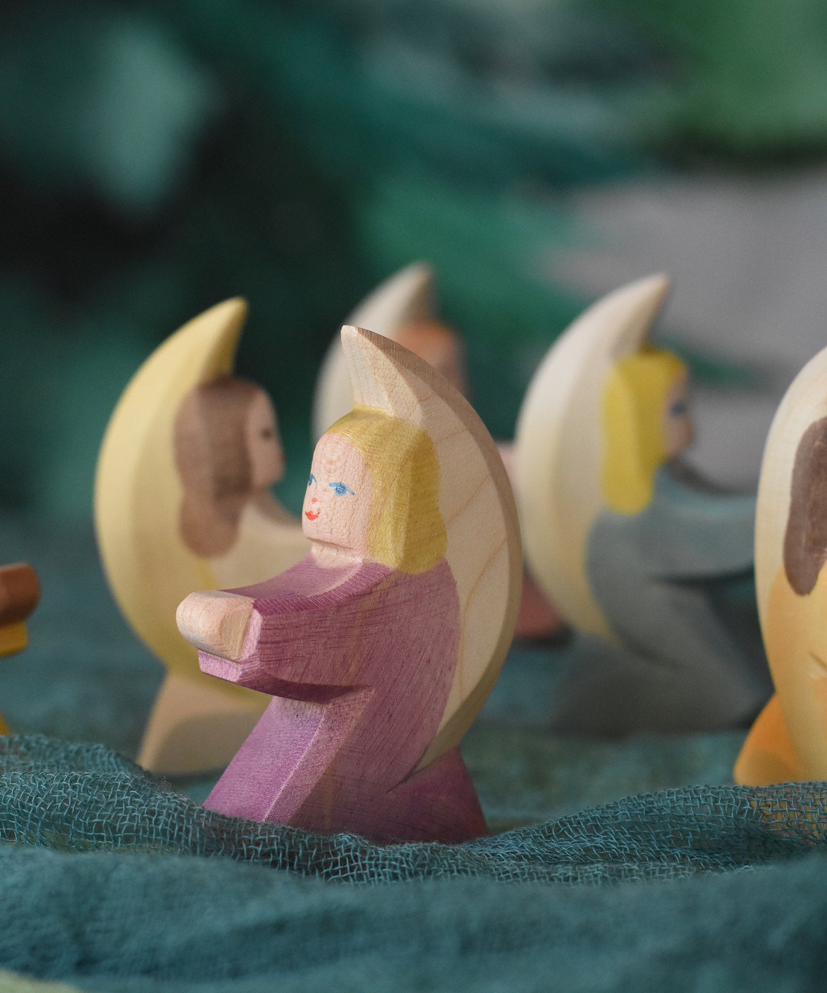 A purple coloured little Ostheimer angel placed on a green decoration scarf in a small world play set up. Other little angels can be seen in the background. These wooden figures are part of a wide range of expertly hand crafted wooden toys here at Babipur.