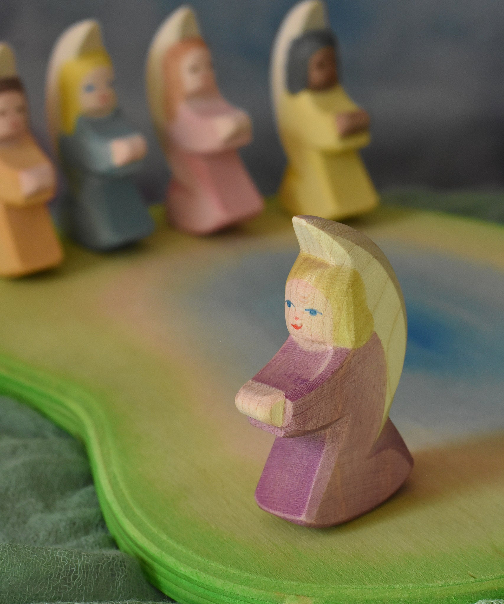 A purple Ostheimer angel placed on an Ostheimer pond scene piece. Other little angels can be seen in the background. This wooden figure is part of a large range of expertly hand crafted, Waldorf inspired, wooden toys here at Babipur.