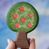 An Ostheimer wooden toy apple tree being held in an adult's hand to show scale. The apple tree has two decorated sides, this side shows the red apples. This wooden figure is part of a large range of expertly hand crafted, Waldorf inspired, wooden toys here at Babipur. 