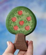 An Ostheimer wooden toy apple tree being held in an adult's hand to show scale. The apple tree has two decorated sides, this side shows the red apples. This wooden figure is part of a large range of expertly hand crafted, Waldorf inspired, wooden toys here at Babipur. 