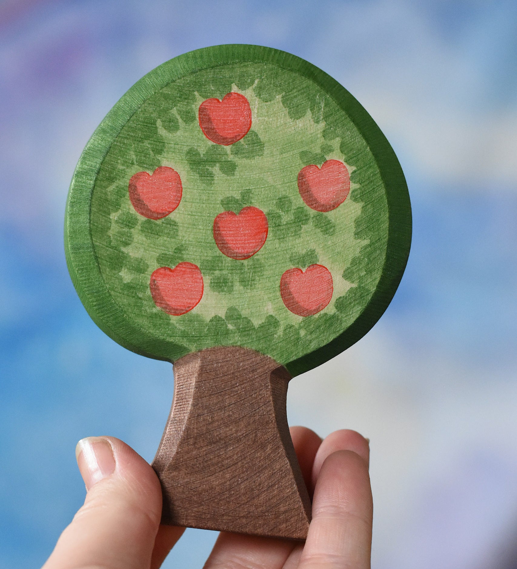 An Ostheimer wooden toy apple tree being held in an adult's hand to show scale. The apple tree has two decorated sides, this side shows the red apples. This wooden figure is part of a large range of expertly hand crafted, Waldorf inspired, wooden toys here at Babipur. 
