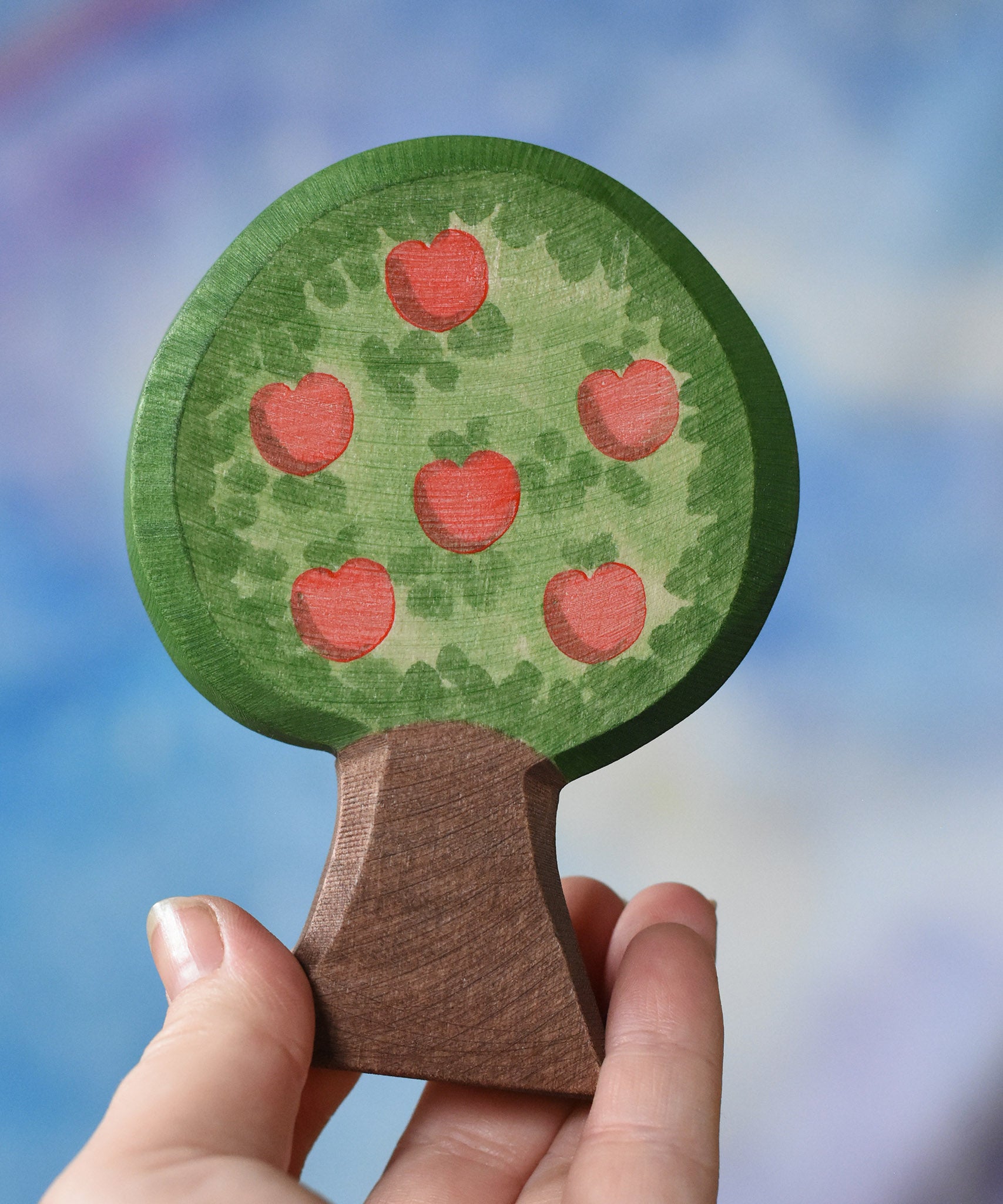An Ostheimer wooden toy apple tree being held in an adult's hand to show scale. The apple tree has two decorated sides, this side shows the red apples. This wooden figure is part of a large range of expertly hand crafted, Waldorf inspired, wooden toys here at Babipur. 
