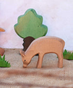An Ostheimer red eating deer shown in a small world play scene with Ostheimer grass an Oak tree seen in the background. Check out our full range of expertly crafted wooden toy figures and scenery here at Babipur.