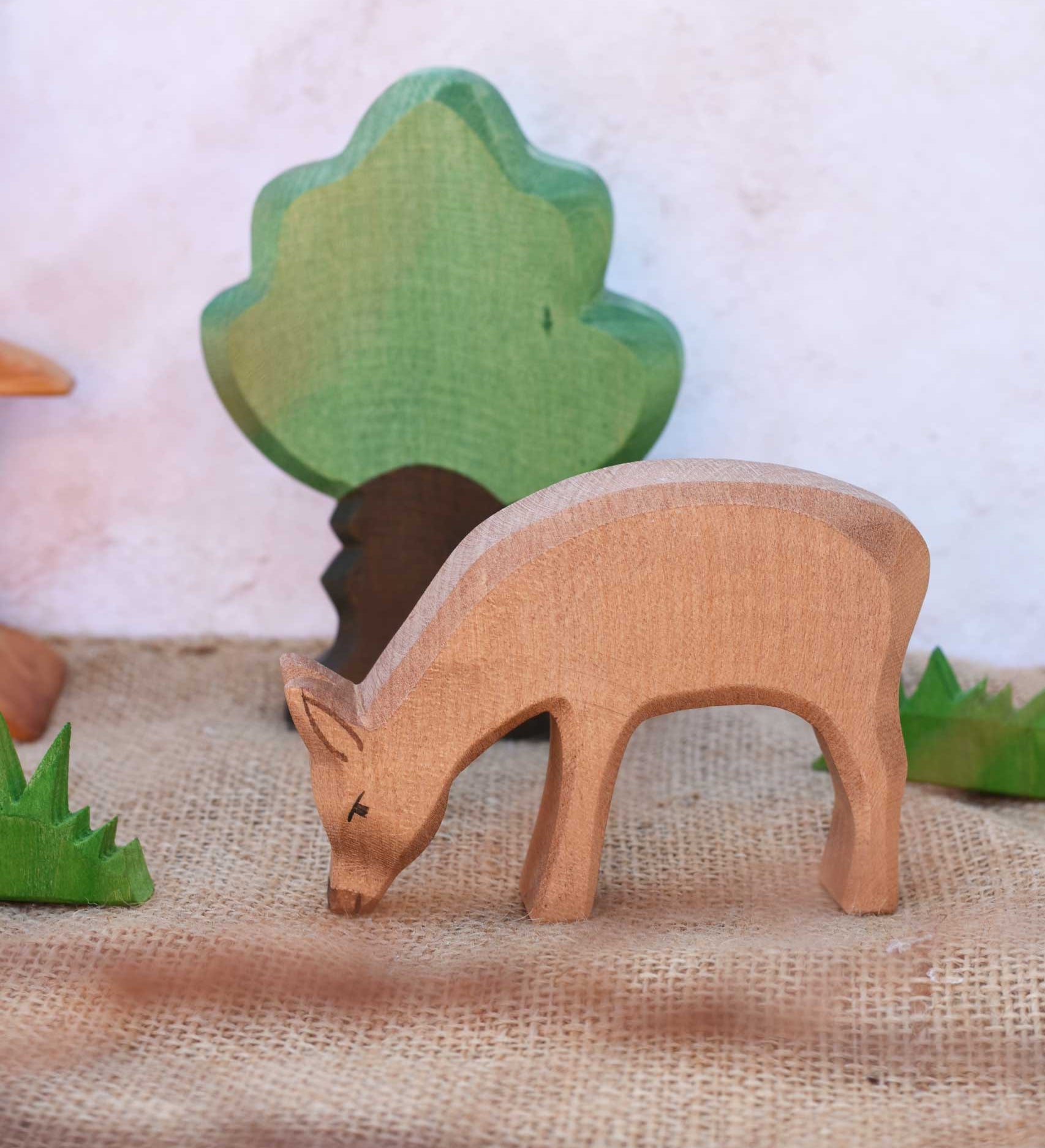 An Ostheimer red eating deer shown in a small world play scene with Ostheimer grass an Oak tree seen in the background. Check out our full range of expertly crafted wooden toy figures and scenery here at Babipur.
