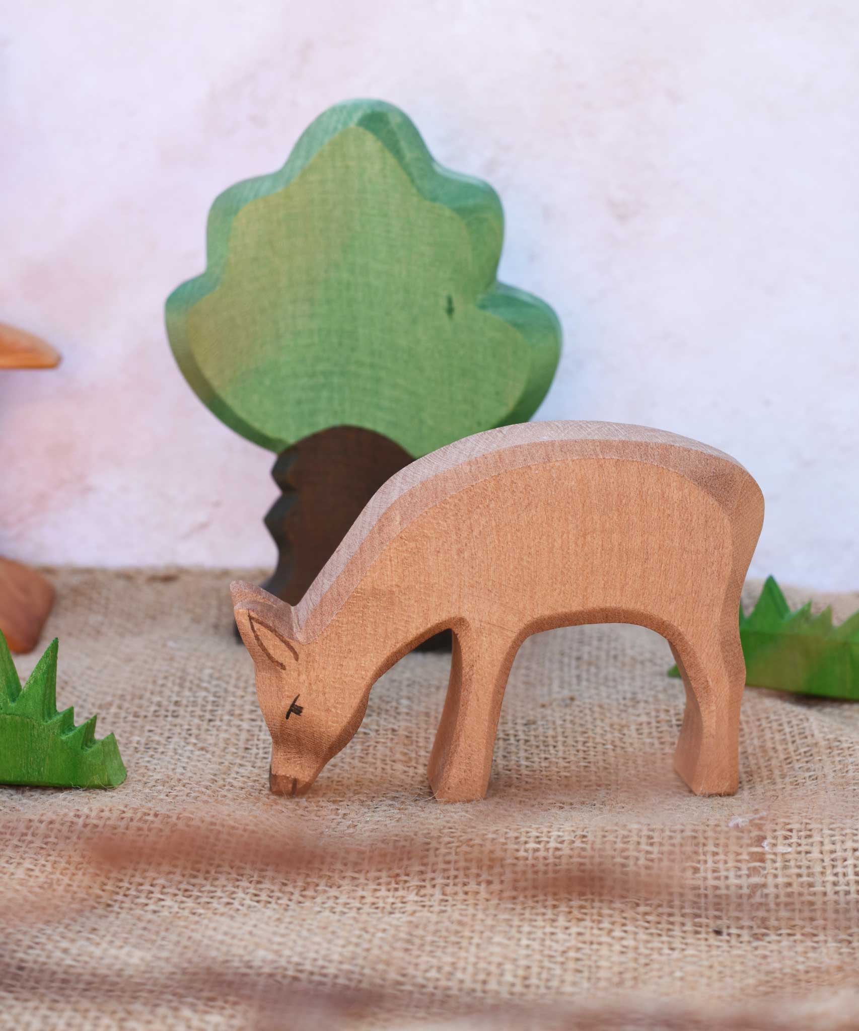 An Ostheimer red eating deer shown in a small world play scene with Ostheimer grass an Oak tree seen in the background. Check out our full range of expertly crafted wooden toy figures and scenery here at Babipur.