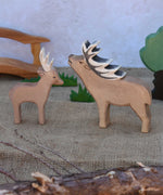An Ostheimer male deer pictured nextto a red deer stag in a small world play scene with Ostheimer grass, oak tree and wooden toy bridge in the background. Check out our full range of expertly crafted wooden toy figures and scenery here at Babipur.
