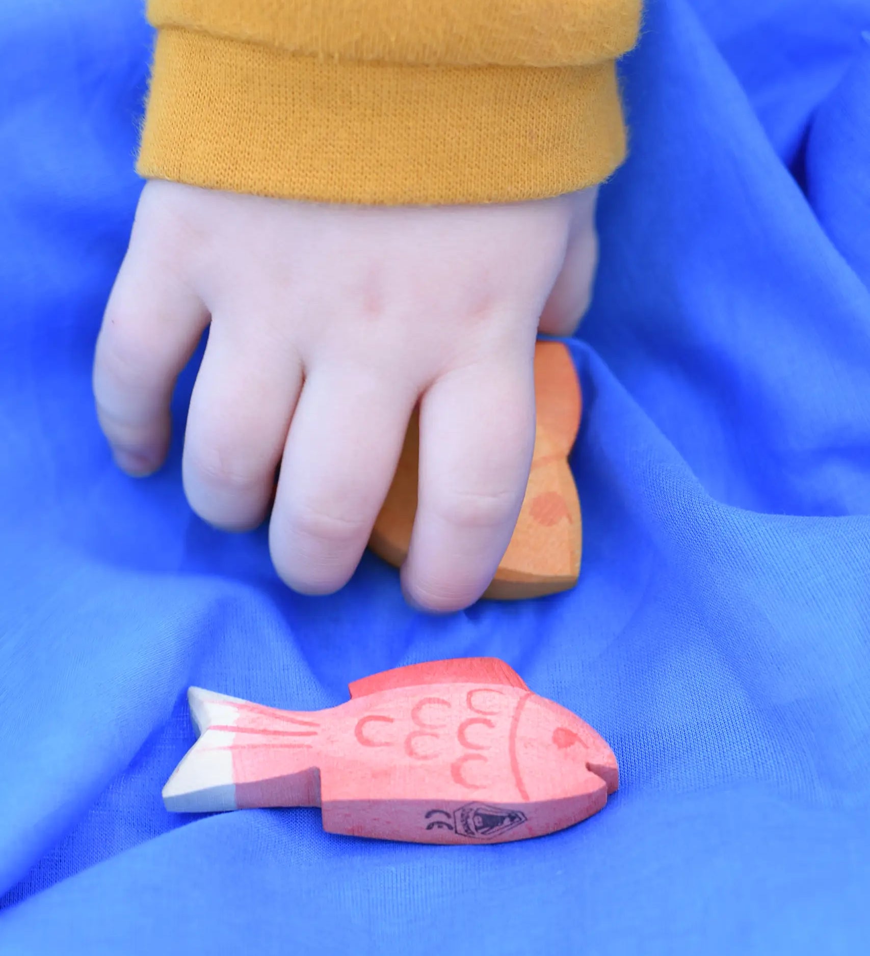 Ostheimer red fish figure placed on a blue Laura's cotton play cloth. A child's hand can be seen behind picking the yellow fish up.