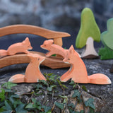 Two Ostheimer sitting fox figures placed at the front of a small world play scene. A wooden toy bridge with small foxes can be seen in the background along with Ostheimer trees and grass. 