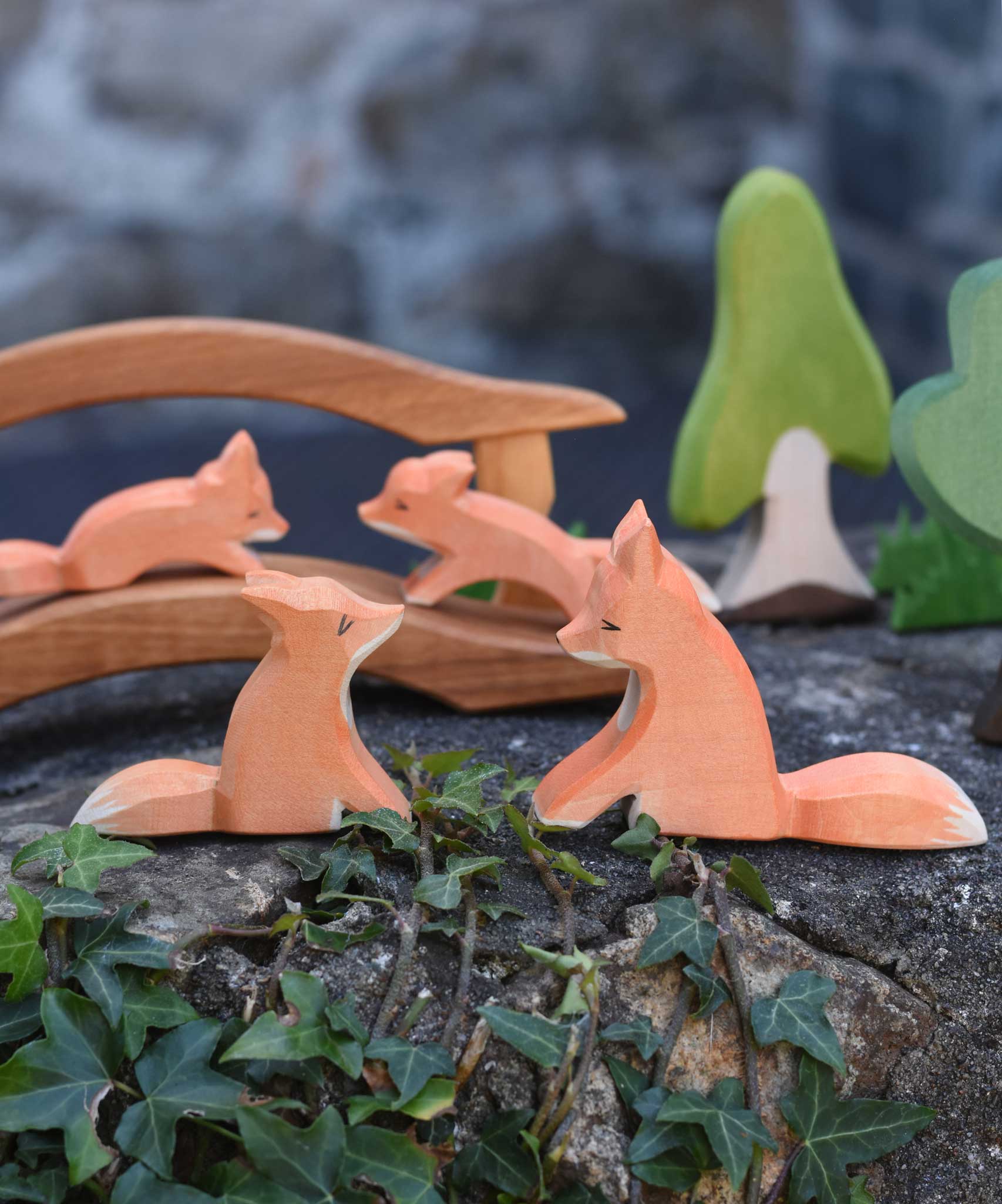 Two Ostheimer sitting fox figures placed at the front of a small world play scene. A wooden toy bridge with small foxes can be seen in the background along with Ostheimer trees and grass. 