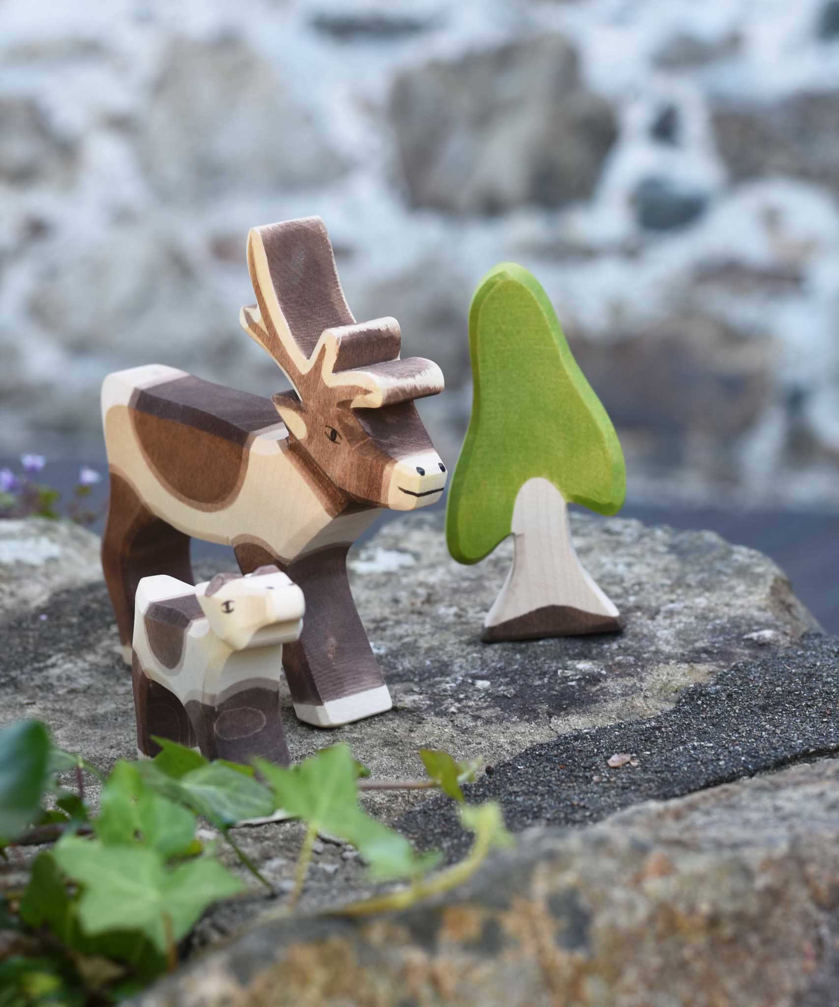 An Ostheimer reindeer and reindeer calf placed on a stone wall outdoors next to an Ostheimer birch tree. Check out our full range of expertly crafted wooden toy figures and scenery here at Babipur.