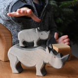 The small Ostheimer rhino wooden toy figure stacked on top of the large rhino. A child's hand can be seen reaching for them in the background. These wooden figures are part of a wide range of expertly hand crafted wooden toys here at Babipur.