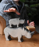 The small Ostheimer rhino wooden toy figure stacked on top of the large rhino. A child's hand can be seen reaching for them in the background. These wooden figures are part of a wide range of expertly hand crafted wooden toys here at Babipur.