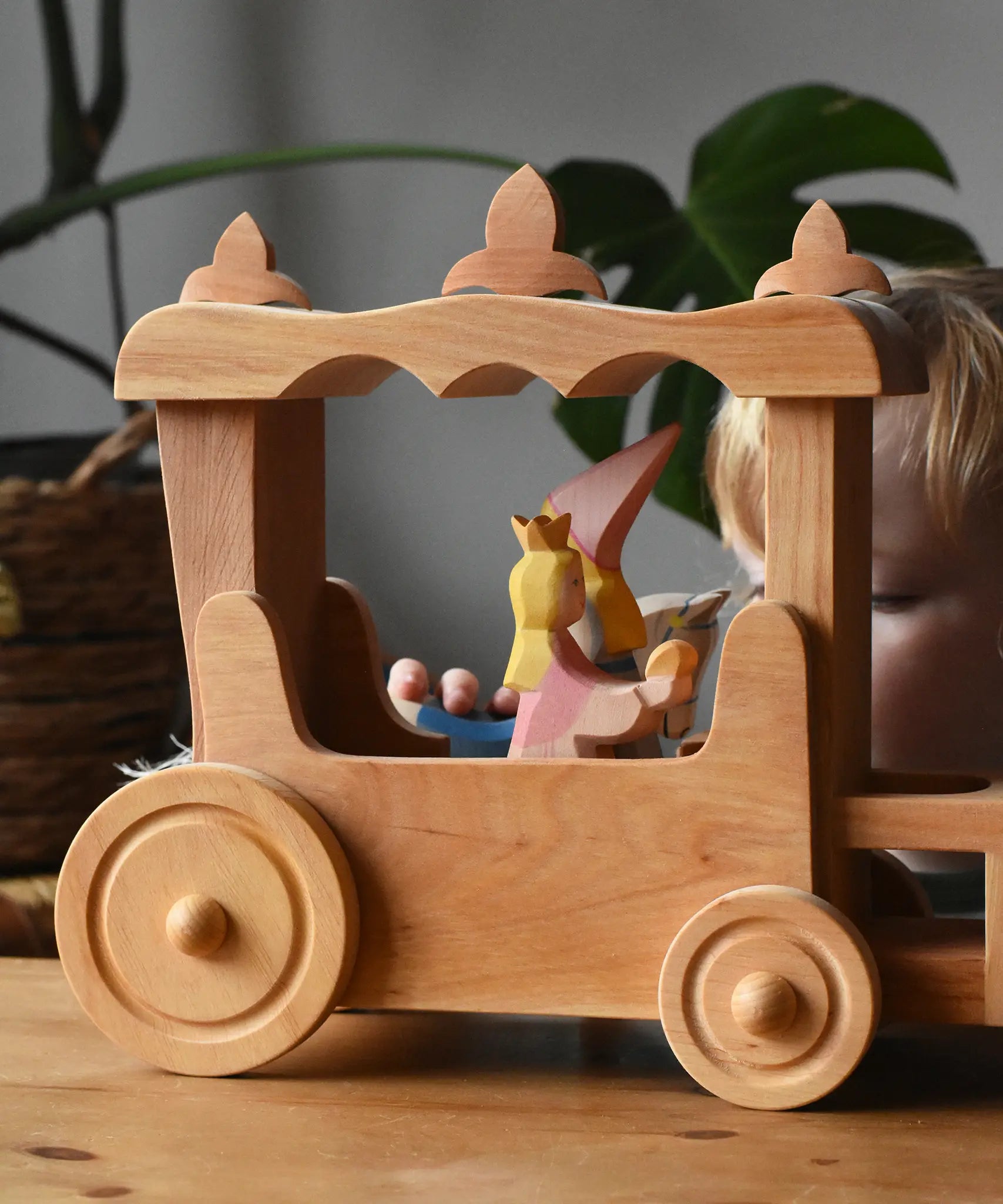 Ostheimer Wooden Stage Coach toy with princess figures placed inside and a child playing in the background.
