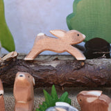 An Ostheimer running rabbit toy figure placed on a log branch in a small world play scene. other Ostheimer rabbits and scenery pieces can be seen in front and in the background. Check out our full range of expertly crafted wooden toy figures and scenery here at Babipur.