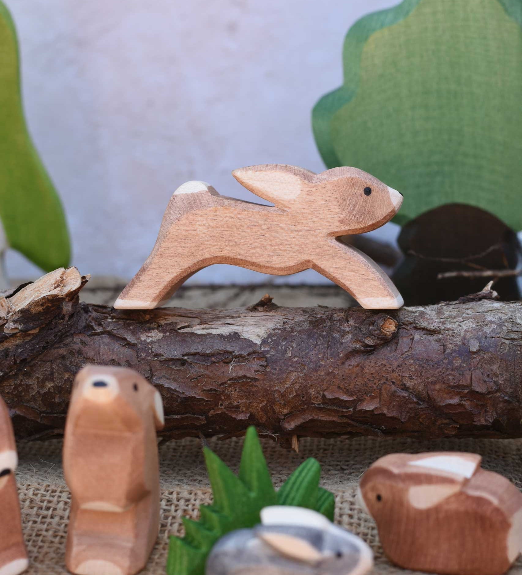 An Ostheimer running rabbit toy figure placed on a log branch in a small world play scene. other Ostheimer rabbits and scenery pieces can be seen in front and in the background. Check out our full range of expertly crafted wooden toy figures and scenery here at Babipur.