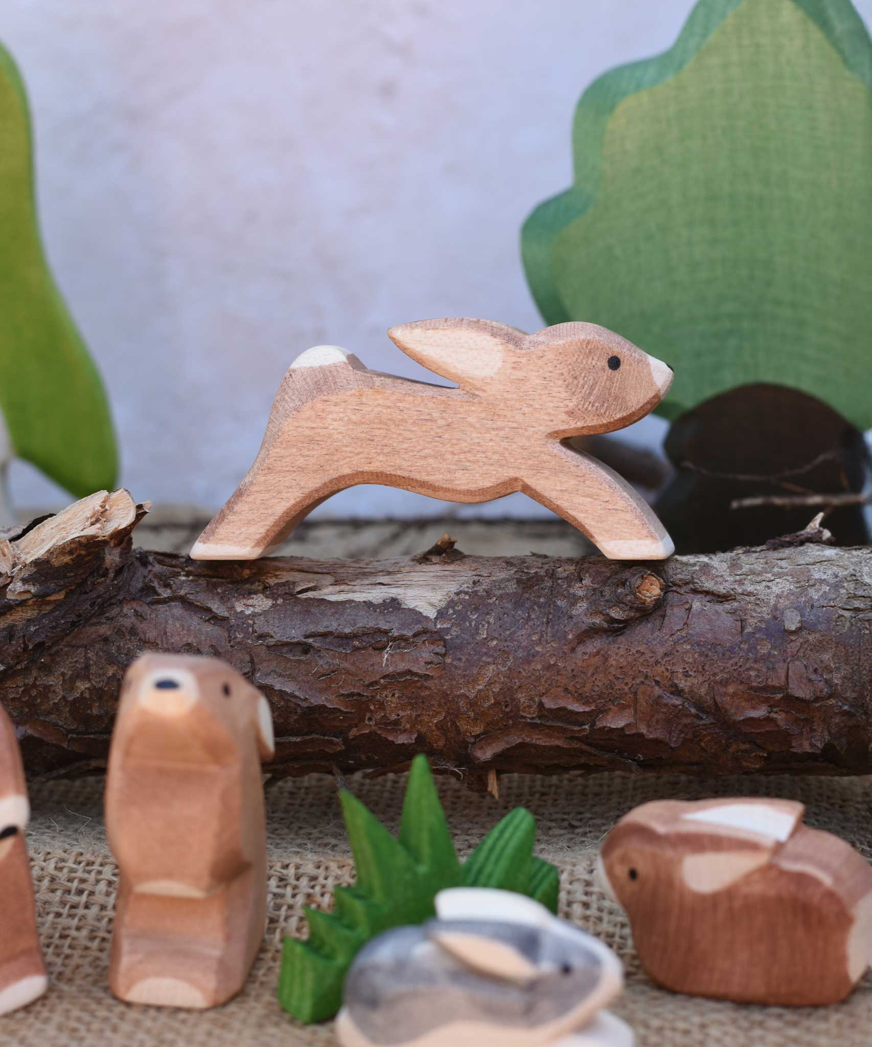 An Ostheimer running rabbit toy figure placed on a log branch in a small world play scene. other Ostheimer rabbits and scenery pieces can be seen in front and in the background. Check out our full range of expertly crafted wooden toy figures and scenery here at Babipur.