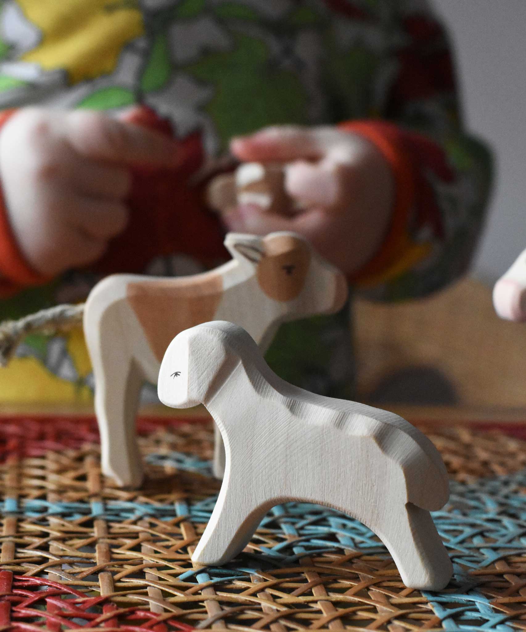 Ostheimer white running lamb wooden toy figure placed at the front of a small world play scene on a rattan top stool.