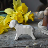 Ostheimer white wooden running lamb figure placed on a wooden surface in front of a bunch of daffodils. 