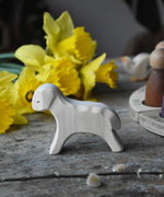 Ostheimer white wooden running lamb figure placed on a wooden surface in front of a bunch of daffodils. 