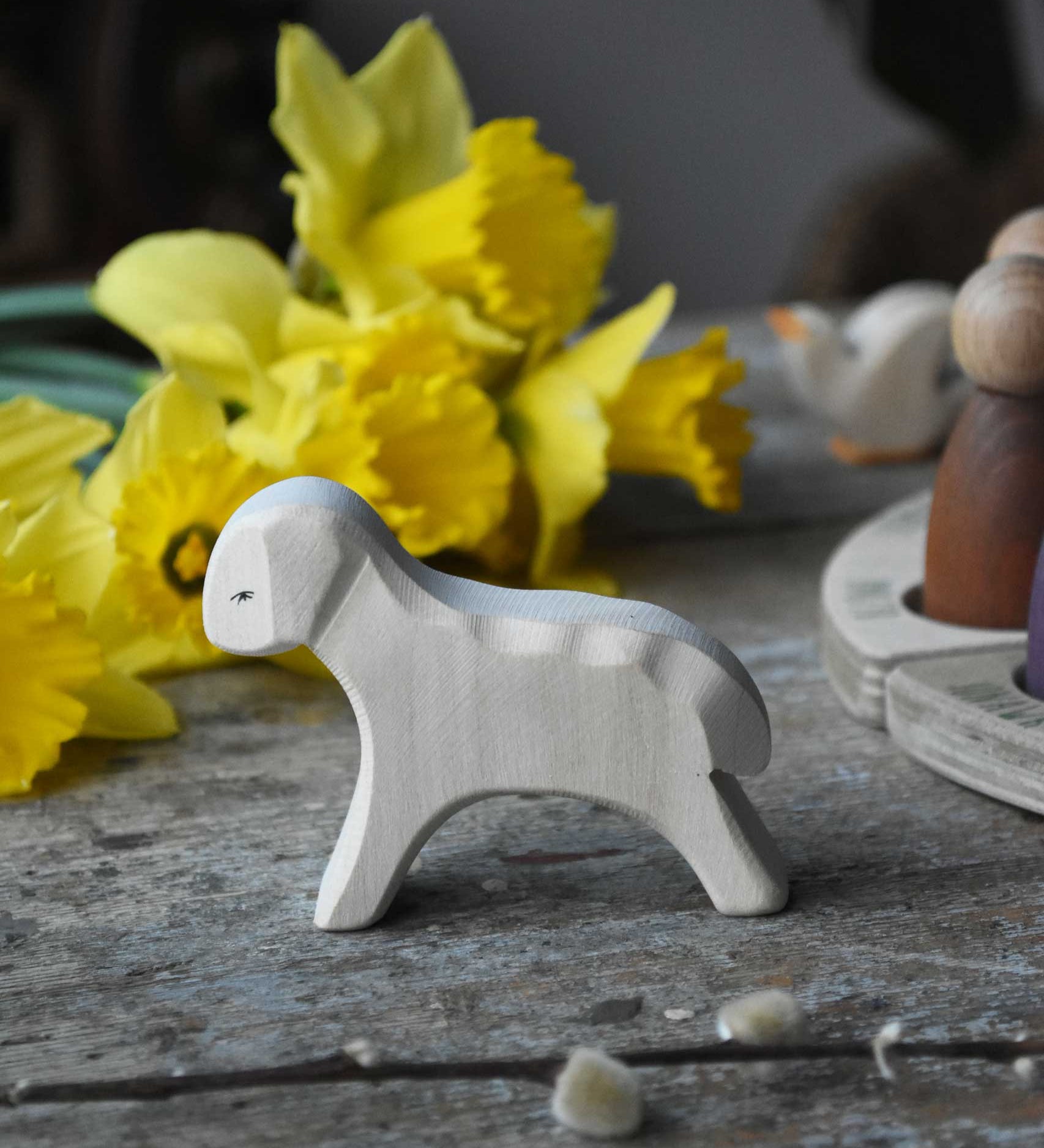 Ostheimer white wooden running lamb figure placed on a wooden surface in front of a bunch of daffodils. 