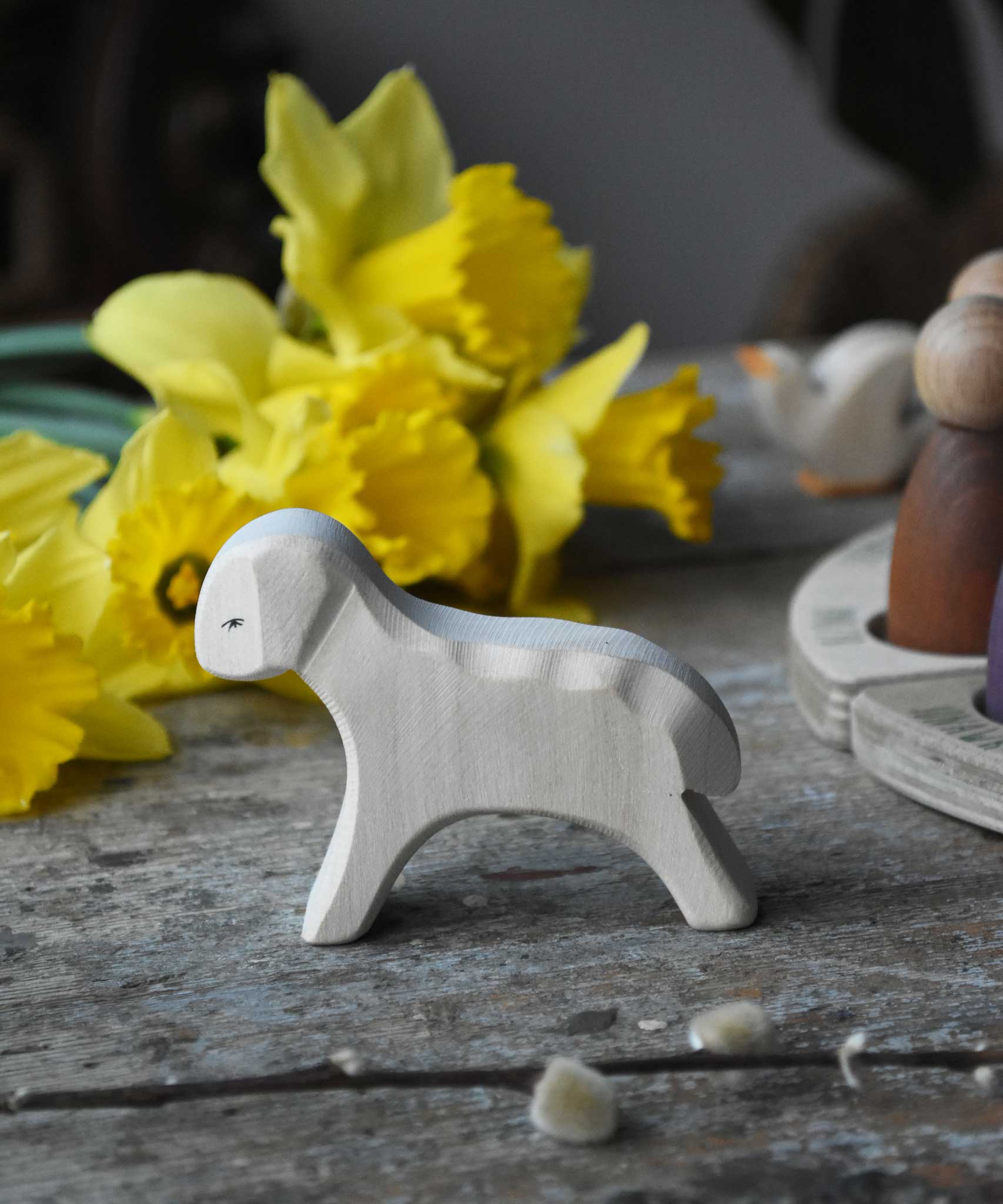Ostheimer white wooden running lamb figure placed on a wooden surface in front of a bunch of daffodils. 