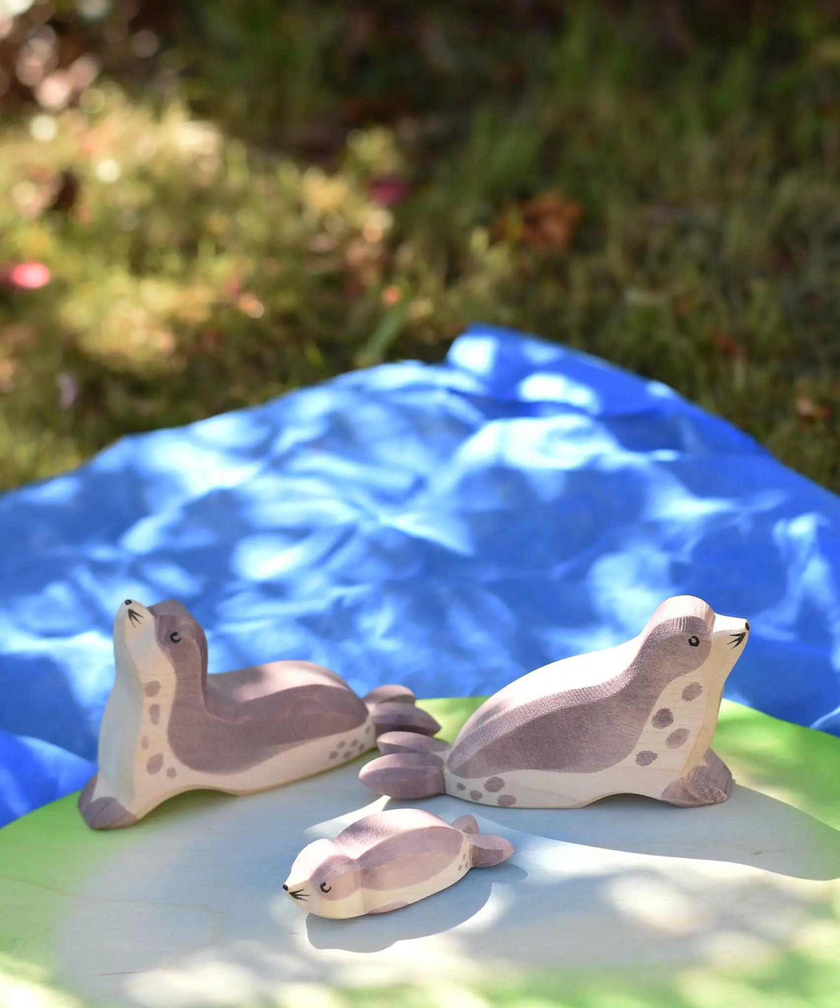 Ostheimer small sea lion pup, head high and head low wooden toy figures placed on a pond piece on blue Laura's cotton play cloth in the Babipur garden.