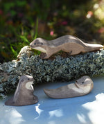 Ostheimer running sea otter wooden toy figure placed on a lichen covered branch with a small swimming and sitting otter placed in front of it. 