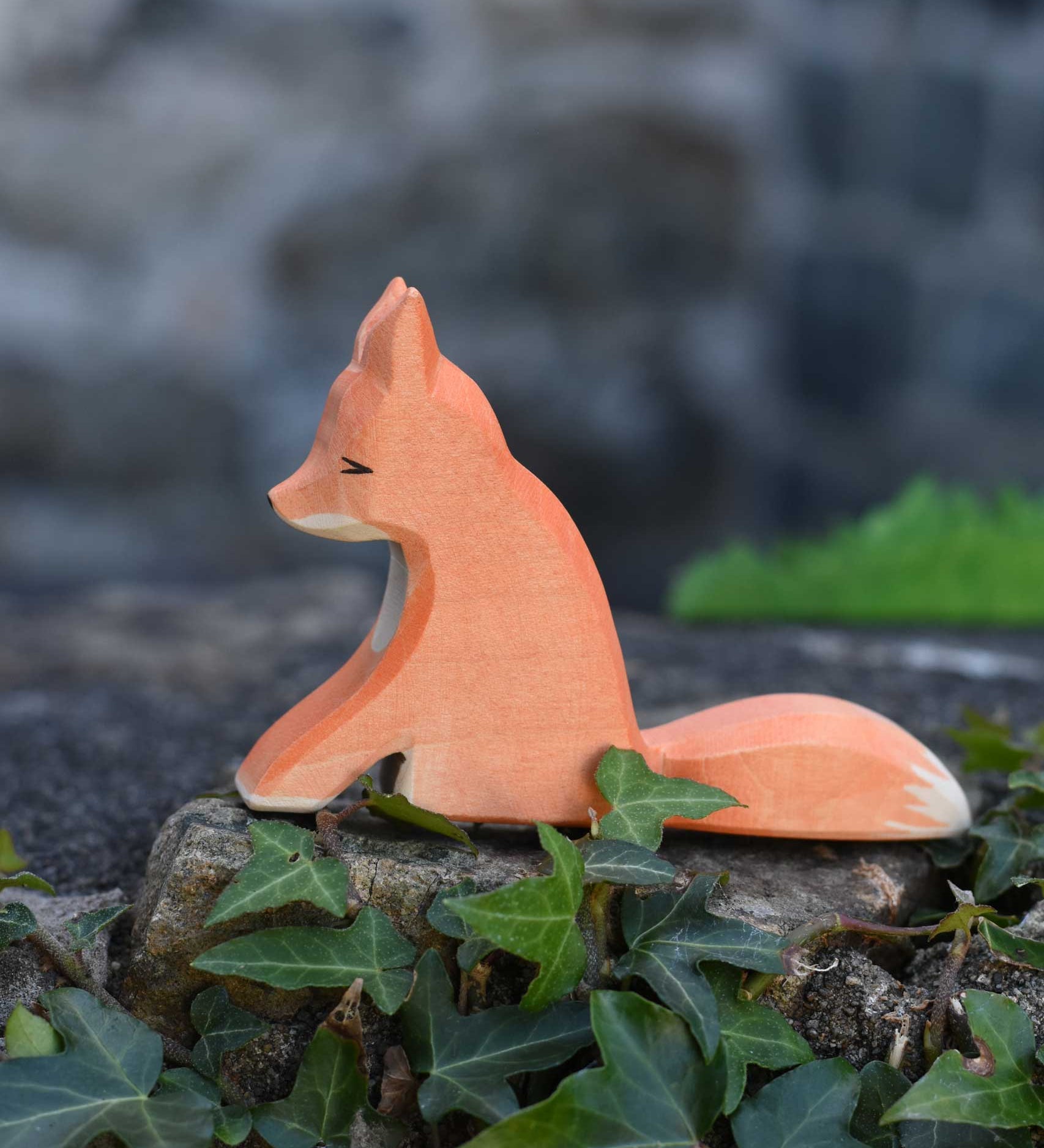 An Ostheimer sitting red fox toy figure placed on a stone wall outdoors. Check out our full range of expertly crafted wooden toy figures and scenery here at Babipur.