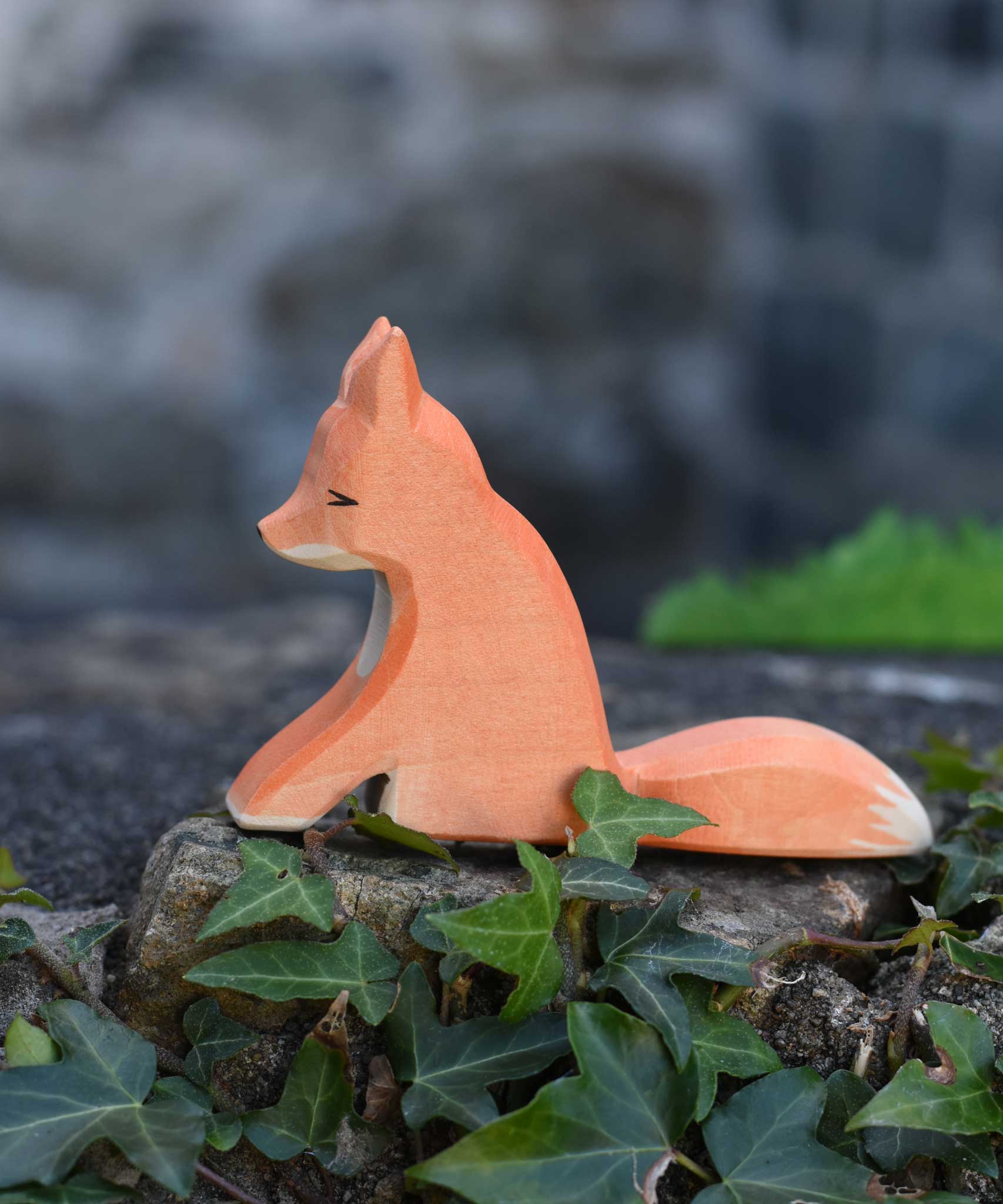 An Ostheimer sitting red fox toy figure placed on a stone wall outdoors. Check out our full range of expertly crafted wooden toy figures and scenery here at Babipur.