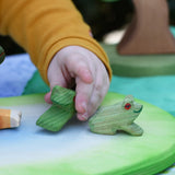 Ostheimer sitting frog handmade wooden toy figure placed on a pond play piece, child in the background playing with the jumping frog toy.
