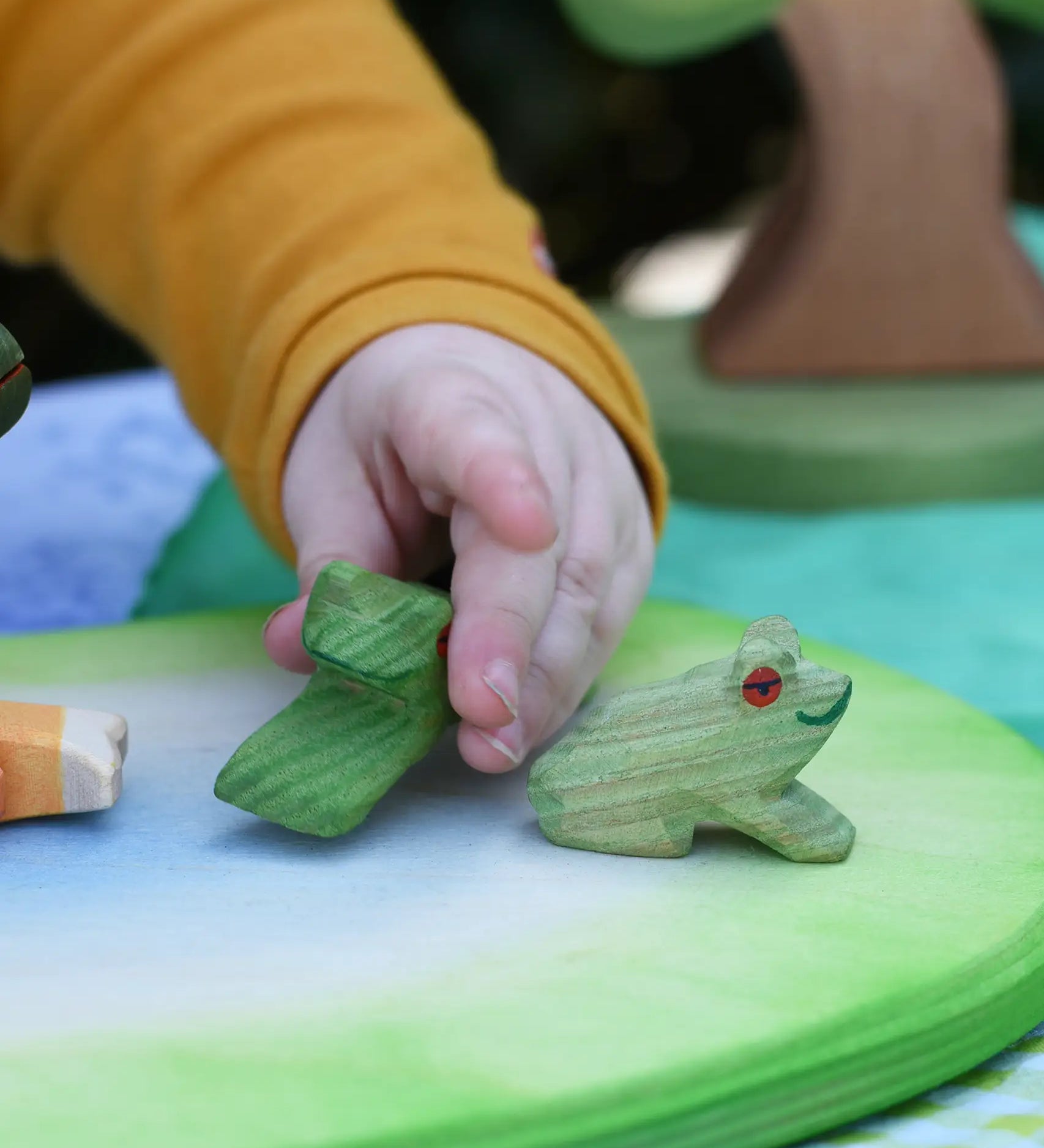 Ostheimer sitting frog handmade wooden toy figure placed on a pond play piece, child in the background playing with the jumping frog toy.