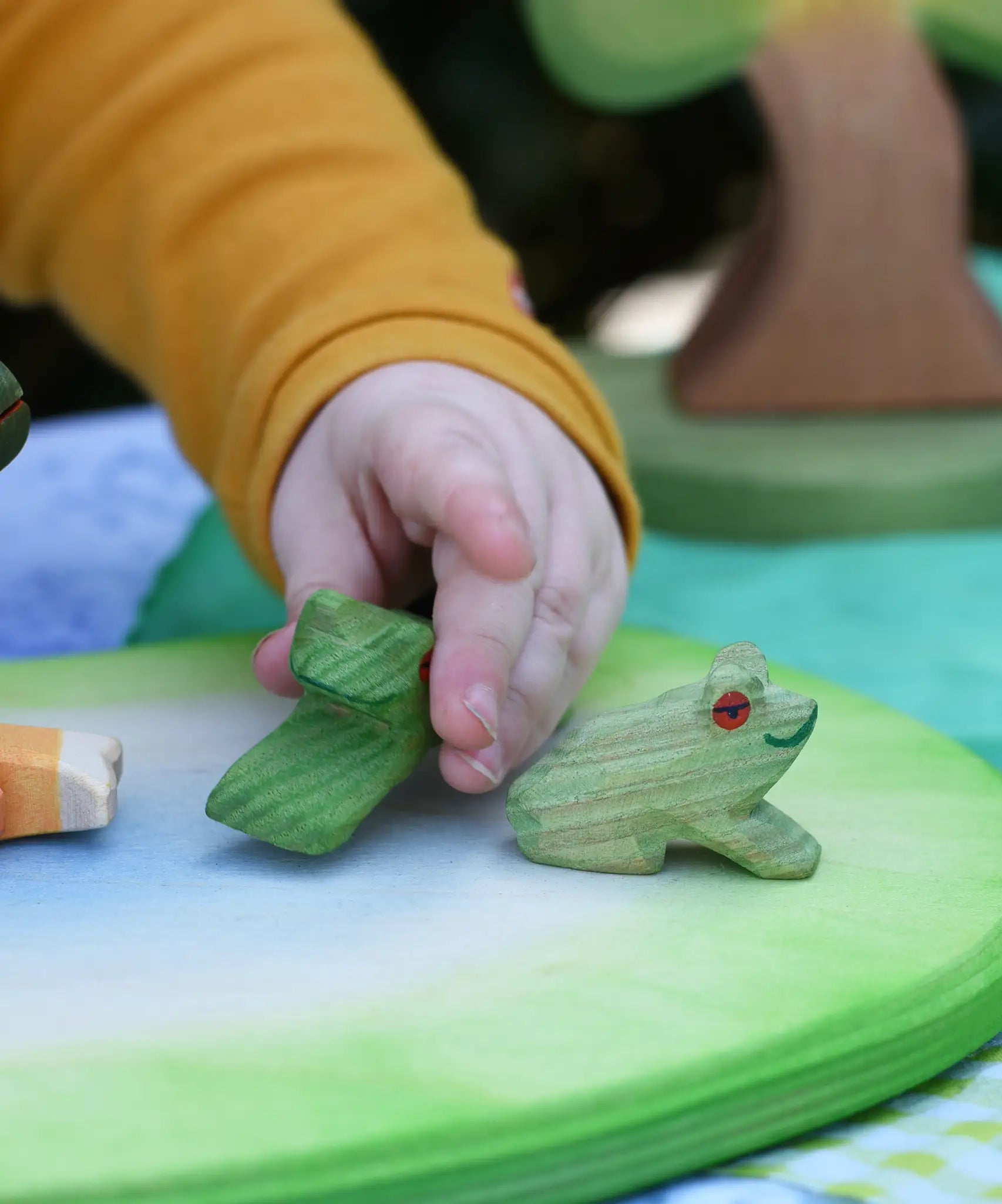 Ostheimer sitting frog handmade wooden toy figure placed on a pond play piece, child in the background playing with the jumping frog toy.