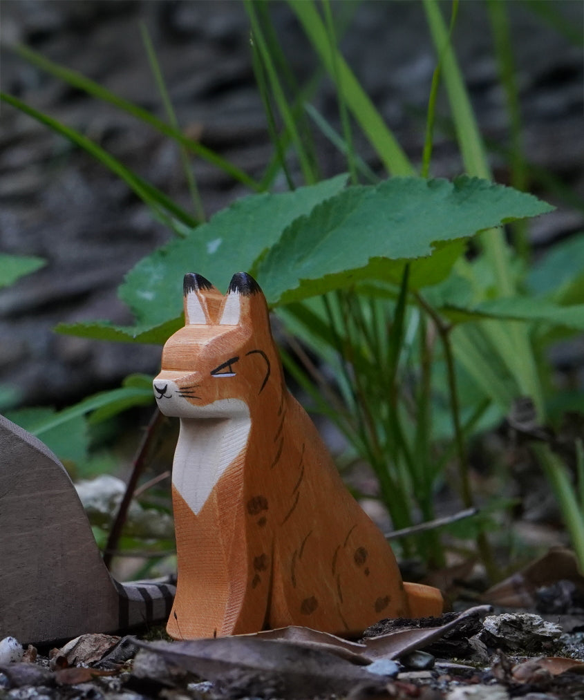 Ostheimer Wooden Sitting Lynx figure placed in front of grass and greenery outdoors. This expertly hand crafted wooden figure is part of a large range of wooden toys here at Babipur.
