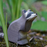 An Ostheimer sitting raccoon wooden figure placed on a mossy surface outdoors among blades of green grass. This expertly hand crafted wooden figure is part of a large range of wooden toys here at Babipur.