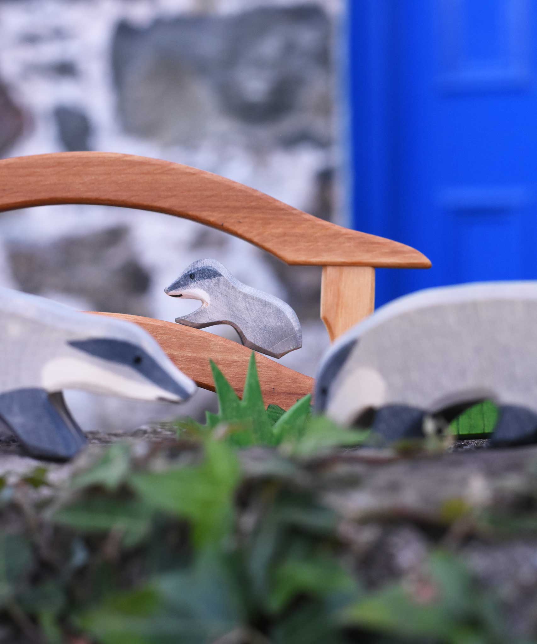 A small Ostheimer badger figures placed on a wooden toy bridge in a small world play scene. Two bigger badgers can be seen blurred in the foreground of the photo. 