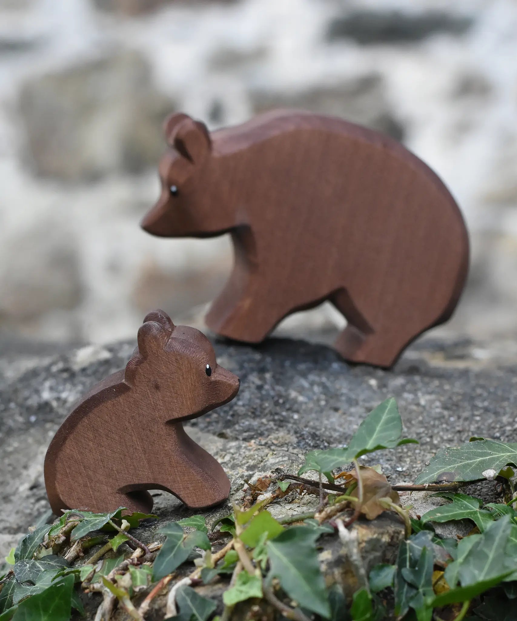 Ostheimer small sitting brown bear wooden toy figure placed on a stone wall outdoors with the large bear blurred in the background. 