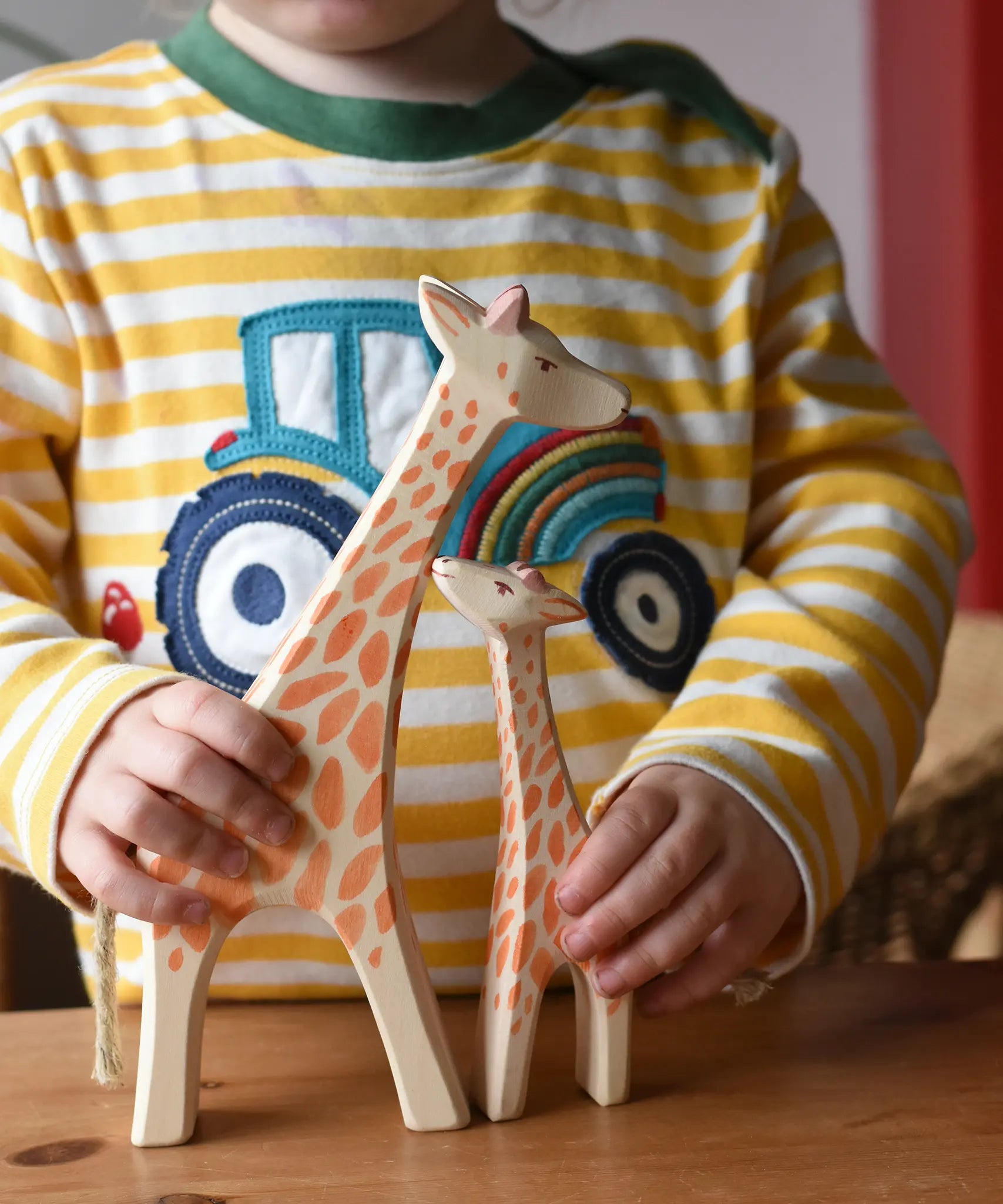 Child holding Ostheimer giraffe wooden toy figures. A running giraffe and small giraffe with head high. 