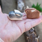Two small Ostheimer rabbit wooden toy figures in an adult's hand to show scale. One rabbit is  Black and White and the other is brown. Check out our full range of expertly crafted wooden toy figures and scenery here at Babipur.