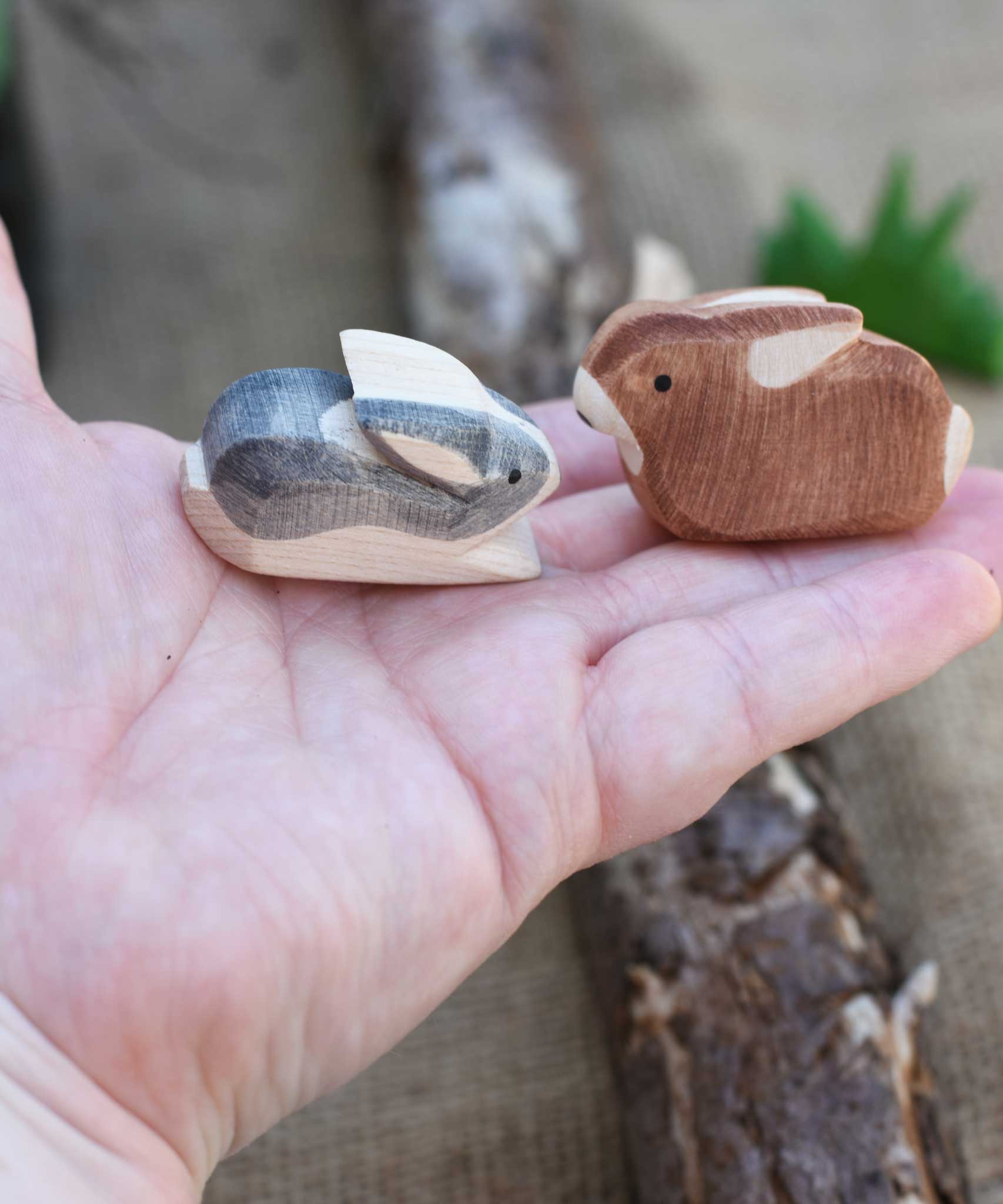 Two small Ostheimer rabbit wooden toy figures in an adult's hand to show scale. One rabbit is  Black and White and the other is brown. Check out our full range of expertly crafted wooden toy figures and scenery here at Babipur.