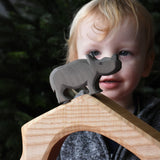 The small Ostheimer rhino wooden toy figure placed on a wooden cave shaped piece. A child can be seen looking on in the background. These wooden figures are part of a wide range of expertly hand crafted wooden toys here at Babipur.