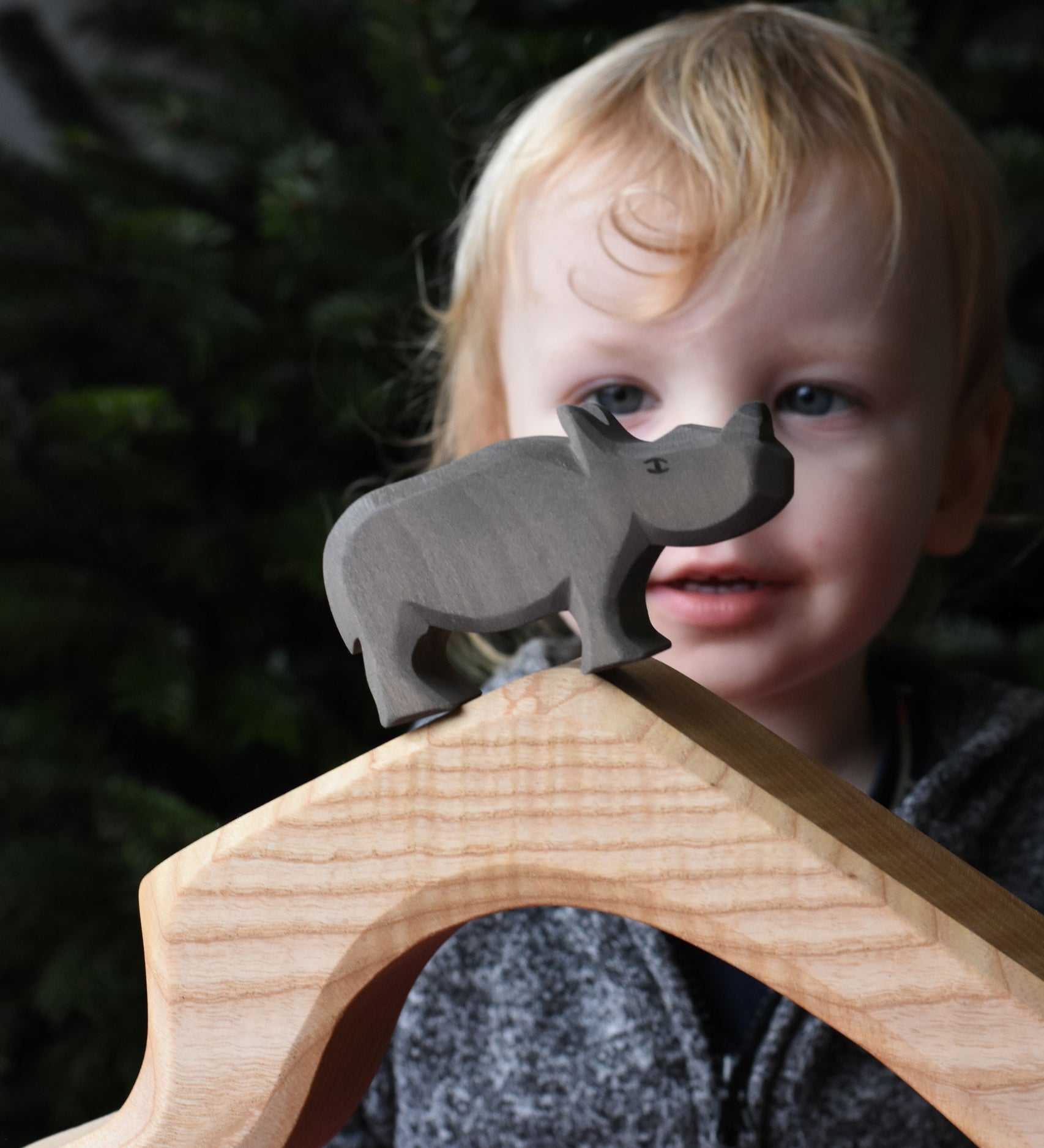 The small Ostheimer rhino wooden toy figure placed on a wooden cave shaped piece. A child can be seen looking on in the background. These wooden figures are part of a wide range of expertly hand crafted wooden toys here at Babipur.