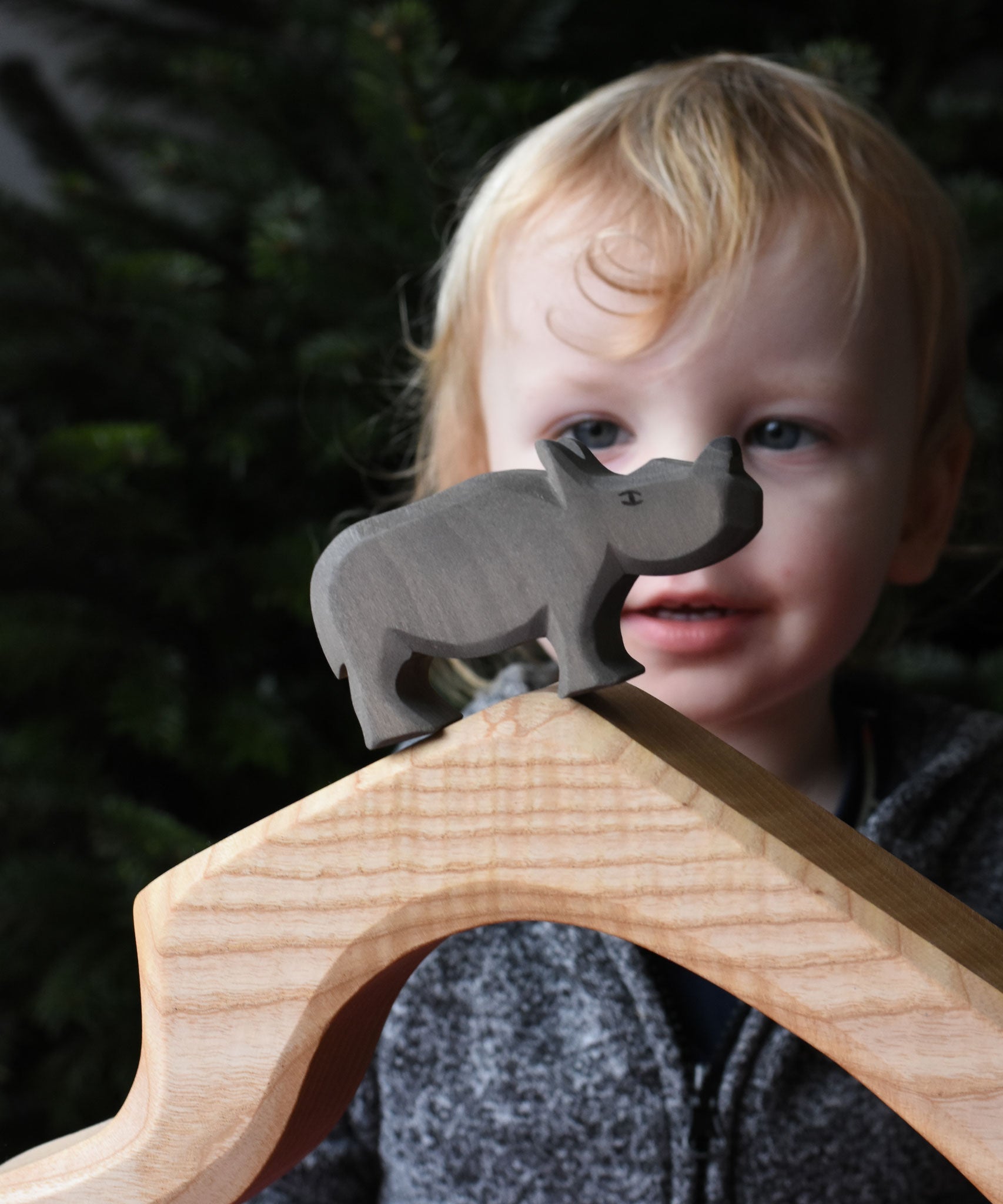 The small Ostheimer rhino wooden toy figure placed on a wooden cave shaped piece. A child can be seen looking on in the background. These wooden figures are part of a wide range of expertly hand crafted wooden toys here at Babipur.