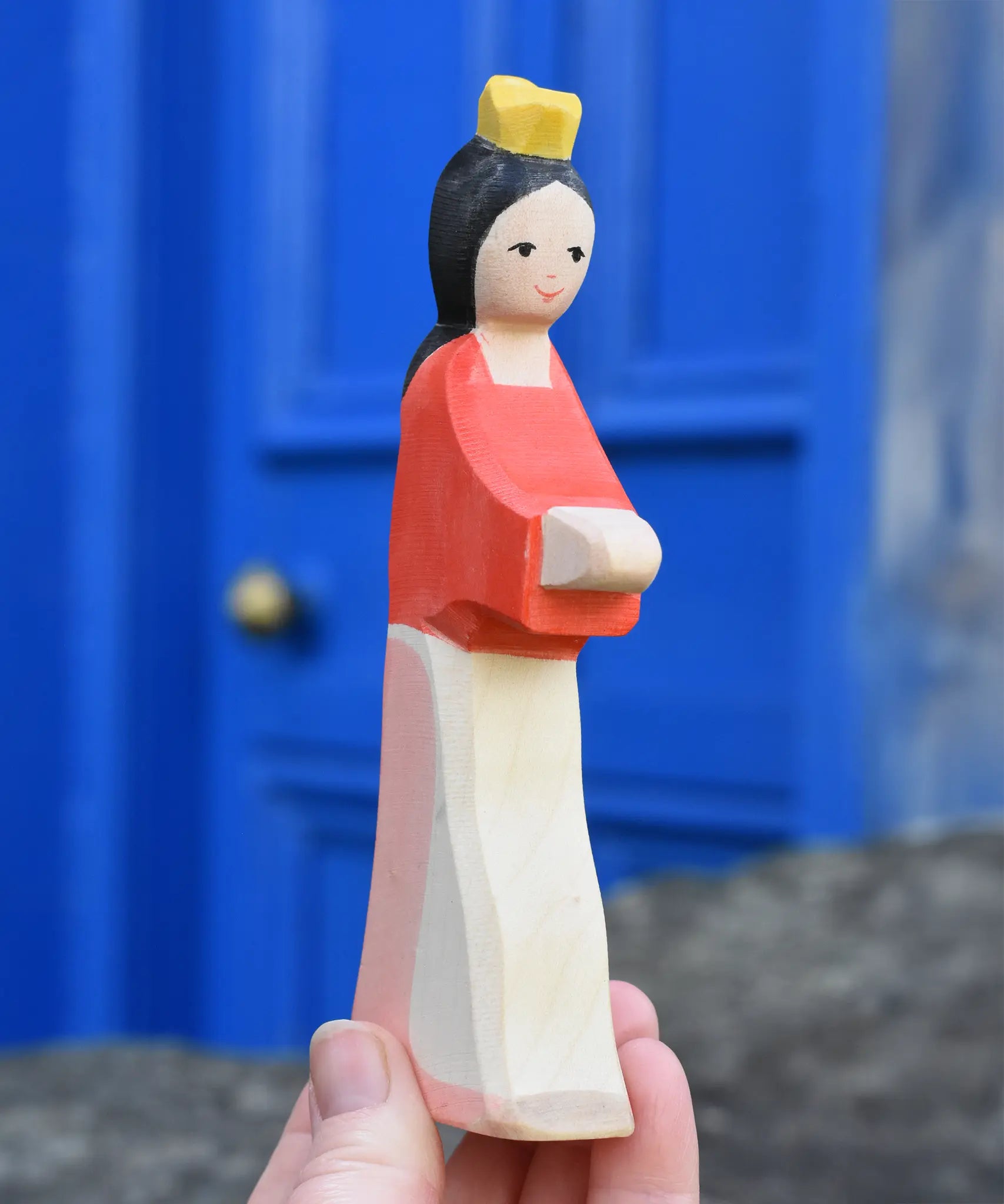 Ostheimer snow white wooden toy figure in an adult's hand held up in front of a blue door.