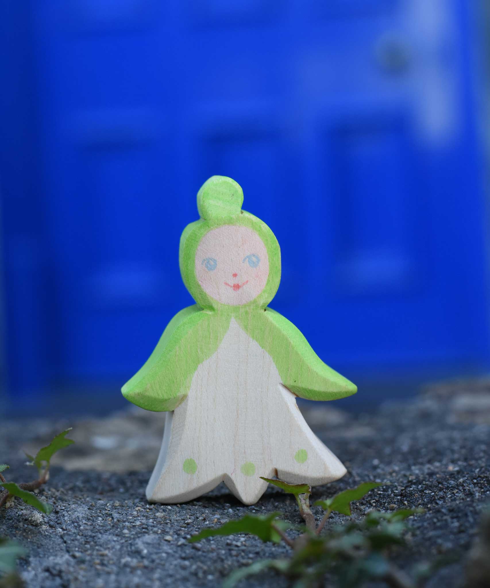 An Ostheimer hand made and hand painted wooden snowdrop Flower Child figure toy placed on a stone wall outdoors. These flower child wooden figures are part of a wide range of expertly hand crafted wooden toys here at Babipur. 