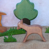 An Ostheimer wooden standing deer figure shown in a small world play scene set up with grass, oak tree and a bridge in the background.