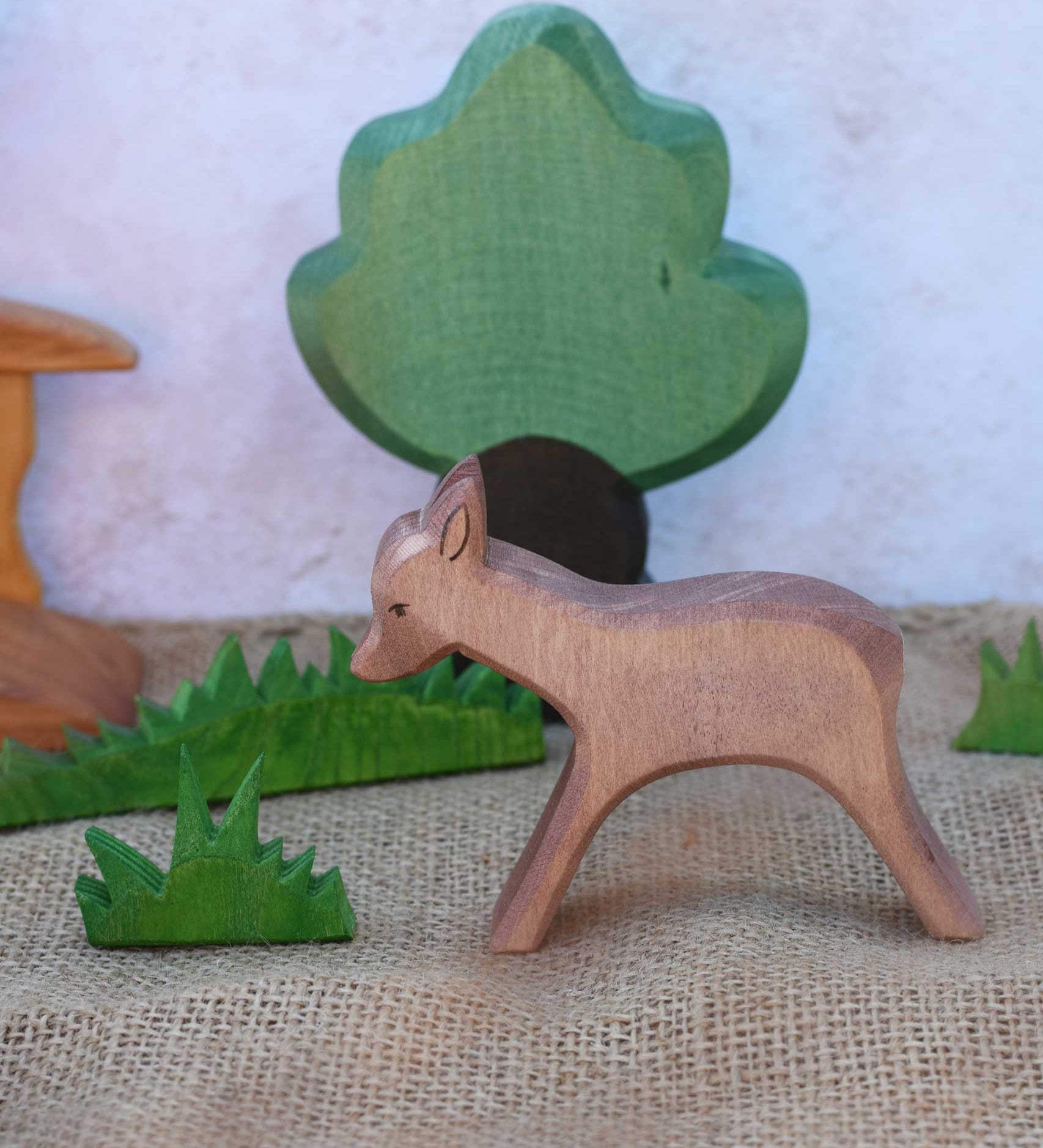 An Ostheimer wooden standing deer figure shown in a small world play scene set up with grass, oak tree and a bridge in the background.